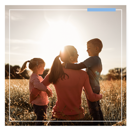 Mom sitting in a field with her arms around her kids. They are all smiling at each other while the sun shines on them.