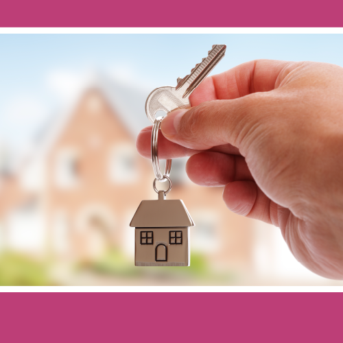 Close up f a person holding a key chain with a house on it. In the background is a blurred house.